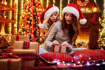 Cheerful mom and her cute daughter girl in santas hats and pajamas exchanging gifts. Parent and little children having fun near tree indoors. Loving family with presents in christmas room.