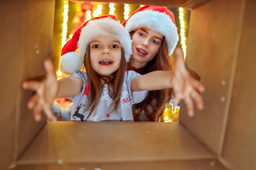 Cheerful mom and her cute daughter girl opening a Christmas present. Parent and little child having fun near Christmas tree indoors. View from inside of the box.