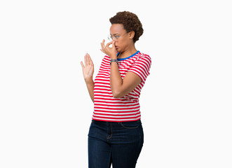 Beautiful young african american woman wearing glasses over isolated background smelling something stinky and disgusting, intolerable smell, holding breath with fingers on nose. Bad smells concept.