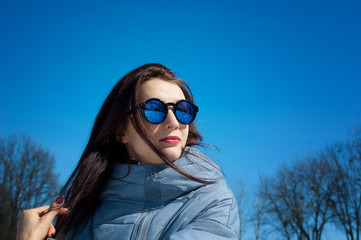 Outdoors lifestyle close up portrait of beautiful girl walking in the snowy winter park. Smiling and enjoying wintertime. Wearing stylish mirrored sunglasses, blue trench coat