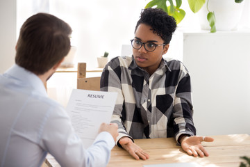 Confused African American millennial female applicant shrug shoulder have misunderstanding with employer or HR manager, businessman consider black candidate resume accuse or blame her for mistake