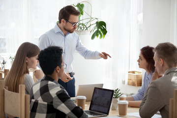 Serious millennial businessman standing leading office meeting talking to female colleague, male...