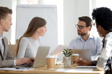 Millennial diverse employees talk brainstorming or collaborating at office meeting, workers sit at shared table with laptops cooperating at casual briefing, colleagues analyze statistics or paperwork