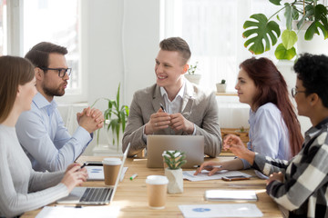Male employee talking at casual company briefing in office, colleagues laugh having fun at meeting with manager, smiling diverse workers brainstorm discussing work issues, joking at workplace