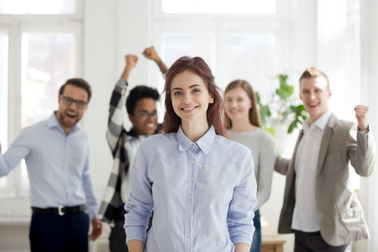 Portrait of smiling female employee standing foreground, excited team or colleagues cheering at background, successful woman professional look at camera posing in office. Leadership concept