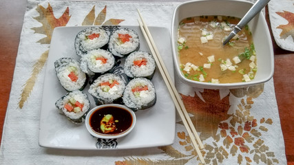 Sushi wrapped in seaweed and filled with smoked salmon, cream cheese and cucumber, with small dipping bowl of soy sauce and wasabi, next to bowl of miso soup with chopsticks on plate