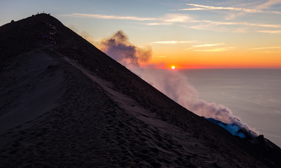 Summit of mount Stromboli at sunset with smoke, a group of tourists and the Mediterranean Sea in...