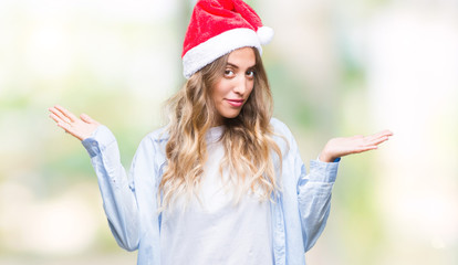 Beautiful young blonde woman wearing christmas hat over isolated background clueless and confused expression with arms and hands raised. Doubt concept.