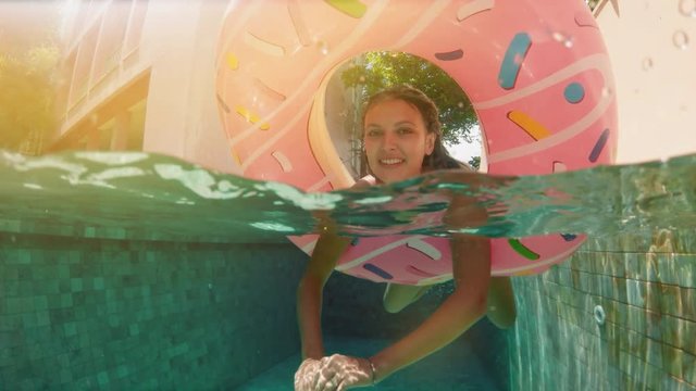 Young hipster millennial girl in sprinkled donut float at pool, smiling look at camera. Young happy woman relaxing on inflatable pool toy in blue swimming pool on sunny day.