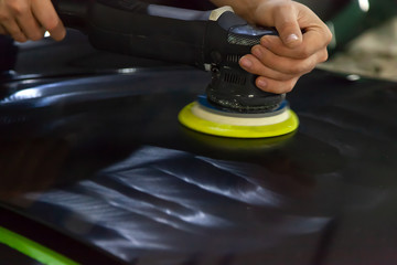 The master in the service station polishes the car body, using a polishing machine, shiny, clean, covered in wax, black hood after painting and washing.