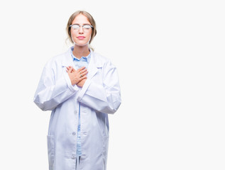 Beautiful young blonde doctor woman wearing white coat over isolated background smiling with hands on chest with closed eyes and grateful gesture on face. Health concept.