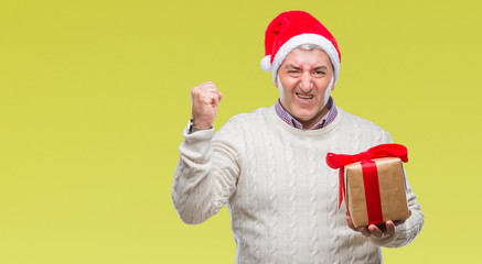 Handsome senior man wearing christmas hat and holding gift over isolated background annoyed and frustrated shouting with anger, crazy and yelling with raised hand, anger concept