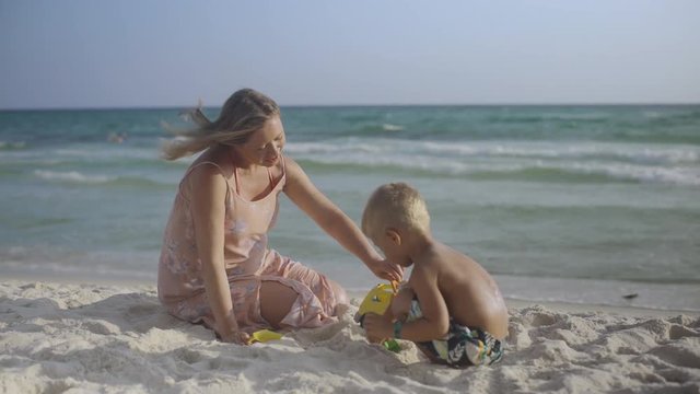 Young pregnant mother sits with her son on the sand on the beach kissing and having fun in summer 4k