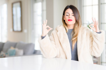 Young beautiful woman wearing winter fur coat at home relax and smiling with eyes closed doing meditation gesture with fingers. Yoga concept.