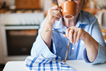 Graceful old hands. Senior woman is drinking hot beverage from mug in cozy home kitchen. Retired person is sitting and holding cup of coffee and spectacles. Grandmother with wedding ring has tea break