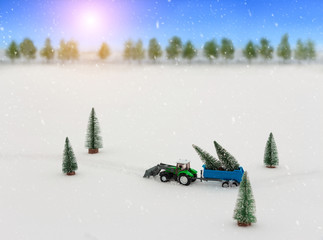 Toy tractor with a trailer carries Christmas trees during snowfall, rides through the snow in the middle of the forest. Beautiful background for greeting card. Winter composition. Happy holiday mood.