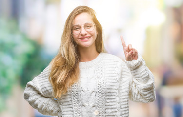 Young beautiful blonde woman wearing winter sweater and sunglasses over isolated background showing and pointing up with finger number one while smiling confident and happy.