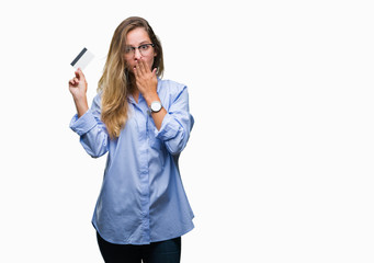 Young beautiful blonde woman holding credit card over isolated background cover mouth with hand shocked with shame for mistake, expression of fear, scared in silence, secret concept