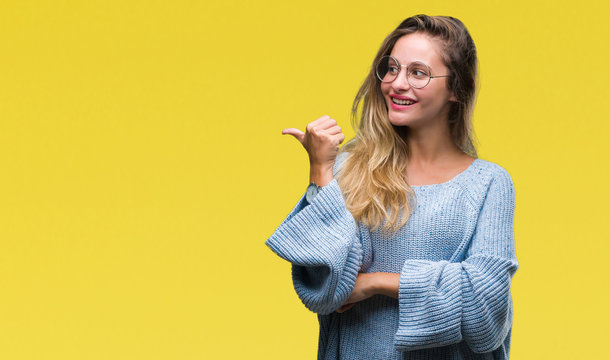 Young beautiful blonde woman wearing sweater and glasses over isolated background smiling with happy face looking and pointing to the side with thumb up.