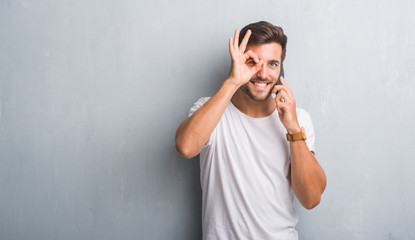 Handsome young man over grey grunge wall speaking on the phone with happy face smiling doing ok sign with hand on eye looking through fingers