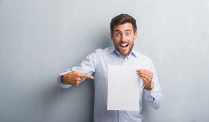 Handsome young man over grey grunge wall holding blank paper sheet contract very happy pointing...
