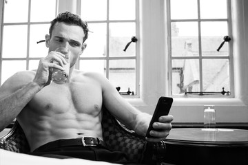 Handsome shirtless man with muscular pecs and six pack abs using mobile phone and drinking juice