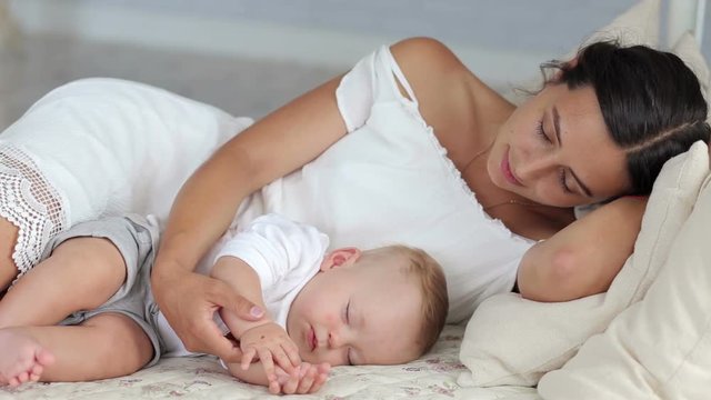 Woman and newborn kid boy relax in a white bedroom. Mom and her newborn baby are sleeping in the bed in the bedroom. Portrait of happy young mom next to sleeping baby in bed.