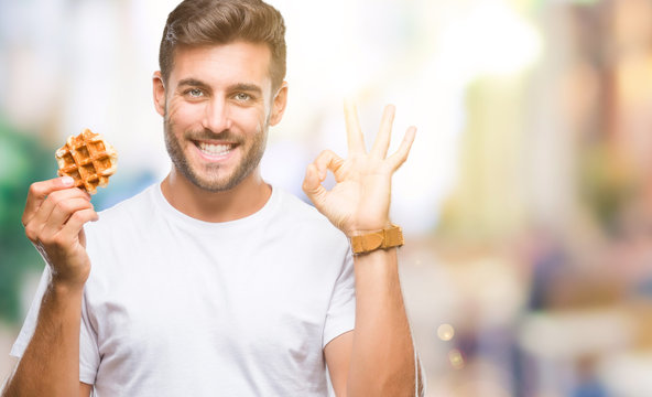 Young handsome man eating a sweet waffle over isolated background doing ok sign with fingers, excellent symbol