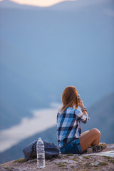 Fototapeta premium Young lady taking selfie with her phone while marveling the view from the top of the world at sunset. Success, winner, zen, tranquility.