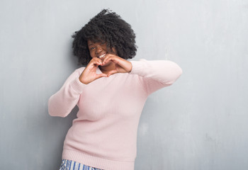 Young african american plus size woman over grey grunge wall wearing winter sweater smiling in love showing heart symbol and shape with hands. Romantic concept.