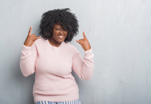 Young african american plus size woman over grey grunge wall wearing winter sweater shouting with crazy expression doing rock symbol with hands up. Music star. Heavy concept.