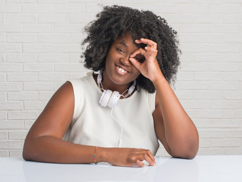 Young african american woman sitting on the table wearing headphones doing ok gesture with hand smiling, eye looking through fingers with happy face.