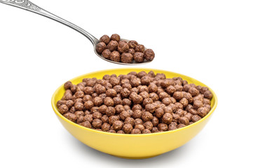 Spoon with chocolate cereal balls over bowl with cereal balls on white.