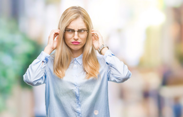 Young beautiful blonde business woman wearing glasses over isolated background covering ears with fingers with annoyed expression for the noise of loud music. Deaf concept.
