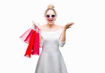 Young beautiful blonde woman holding shopping bags over isolated background very happy and excited, winner expression celebrating victory screaming with big smile and raised hands