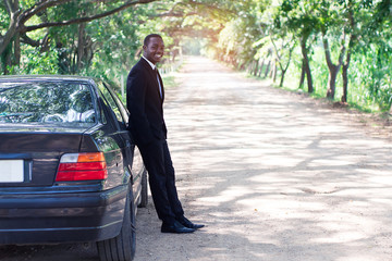 African Businessman in suit standing near car with green natural background.