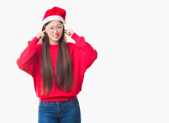 Young Chinese woman over isolated background wearing christmas hat covering ears with fingers with annoyed expression for the noise of loud music. Deaf concept.