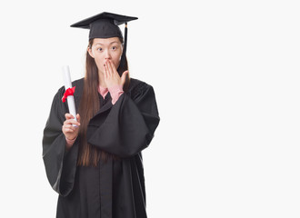 Young Chinese woman wearing graduate uniform holding paper degree cover mouth with hand shocked with shame for mistake, expression of fear, scared in silence, secret concept