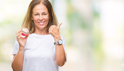 Middle age hispanic woman eating cupcake over isolated background pointing and showing with thumb up to the side with happy face smiling