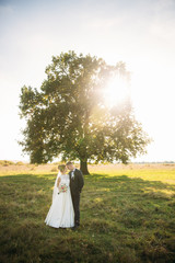 Stylish couple of happy newlyweds walking in field on their wedding day with bouquet. In the middle of the field ther is a big tree