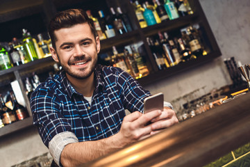 Young bartender leaning on bar counter holding smartphone happy