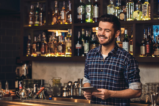 Young bartender standing at bar counter holding digital tablet laughing joyful