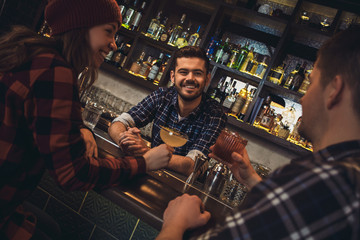 Young bartender standing at bar counter with visitors drinking alcohol talking joyful