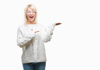 Young beautiful blonde woman wearing winter sweater and glasses over isolated background amazed and smiling to the camera while presenting with hand and pointing with finger.