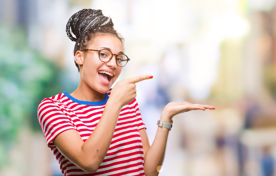 Young braided hair african american girl wearing glasses over isolated background amazed and smiling to the camera while presenting with hand and pointing with finger.