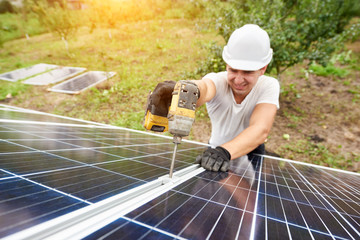 Smiling technician working with screwdriver connecting solar photo voltaic panel to exterior metal platform on sunny green orchard and rural houses background. Efficiency and professionalism concept.