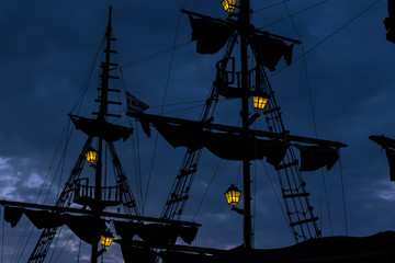 medieval old wooden ship dark mast and sails silhouette at night twilight time on cloudy sky background with many hanging lamps yellow illumination © Artem Kniaz