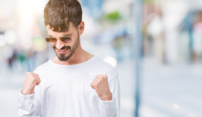 Young handsome man wearing sunglasses over isolated background very happy and excited doing winner gesture with arms raised, smiling and screaming for success. Celebration concept.