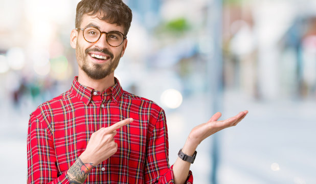 Young handsome man wearing glasses over isolated background amazed and smiling to the camera while presenting with hand and pointing with finger.