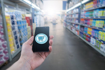 Fototapeta na wymiar Hand holding mobile phone on Supermarket blur background. Shopping basket on a mobile phone screen. 